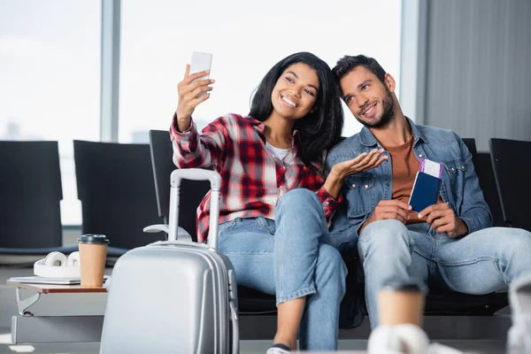 Happy african american woman pointing with hand while taking selfie with bearded man holding passport in airport — Stock Photo