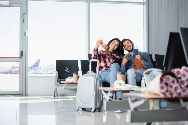 Glücklicher bärtiger Mann zeigt mit dem Finger auf Pass, während er Selfie mit afrikanisch-amerikanischer Frau auf Flughafen macht — Stockfoto