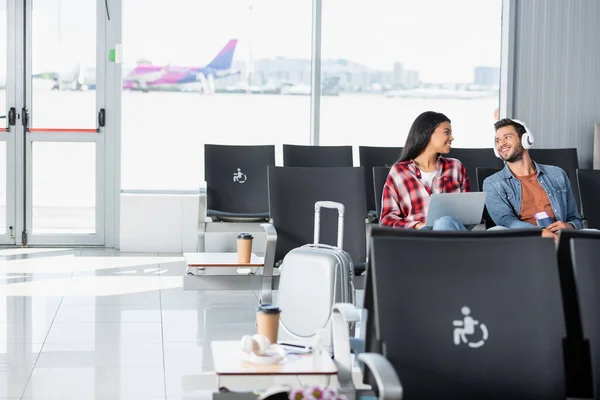 Happy african american woman with laptop looking at bearded man listening music in departure lounge of airport — Stock Photo