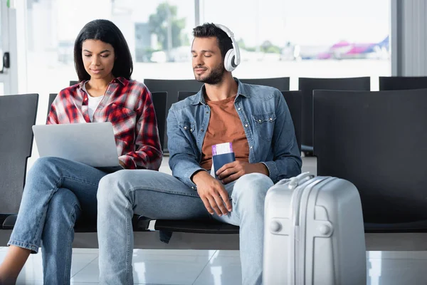 Hombre barbudo escuchando música y mirando portátil cerca de la mujer afroamericana en el aeropuerto - foto de stock