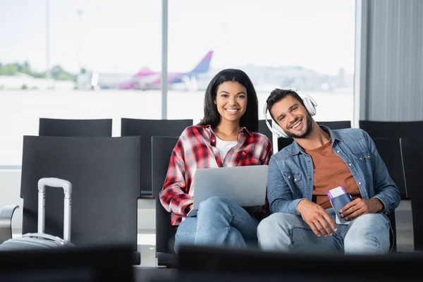 Щасливий чоловік слухає музику біля африканської жінки з ноутбуком в аеропорту. — стокове фото