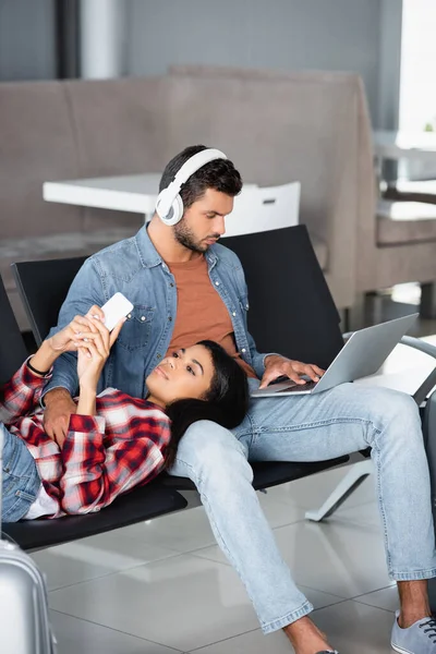 African american woman using phone and lying on bearded man in headphones with laptop in departure lounge — Stock Photo