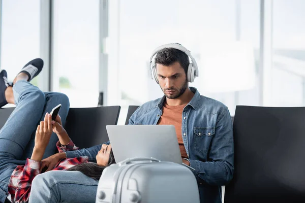 African american woman using phone and lying on bearded man in headphones with laptop in departure lounge — Stock Photo