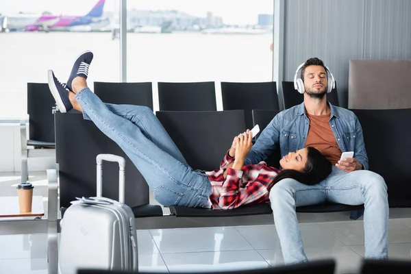 African american woman using phone and lying on bearded man in headphones in departure lounge — Stock Photo