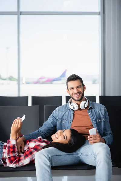 Feliz afroamericana mujer sosteniendo el teléfono y acostado en barbudo hombre sonriendo en la sala de salida — Stock Photo