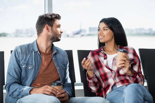 Cheerful multicultural couple holding coffee to go while sitting in departure lounge of airport — Stock Photo