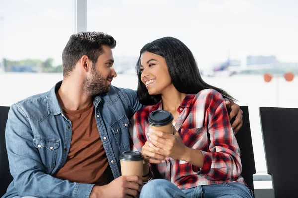 Sourire couple multiculturel tenant un café à emporter assis dans le salon de départ de l'aéroport — Photo de stock