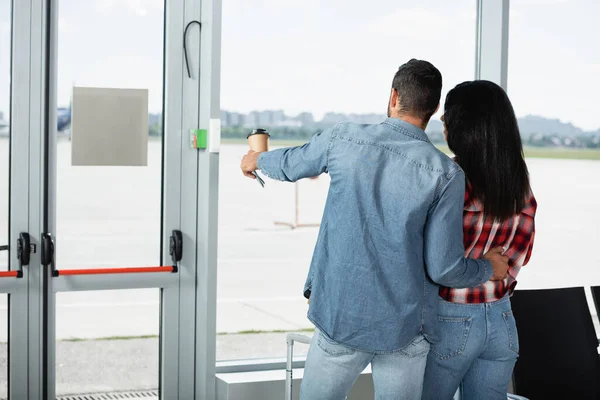 Vue arrière de l'homme tenant café pour aller et pointant vers la femme afro-américaine dans le salon de départ de l'aéroport — Photo de stock
