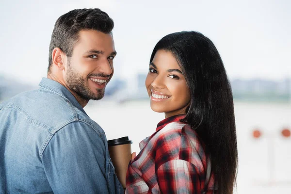 Happy multicultural couple smiling near coffee to go in airport — Stock Photo