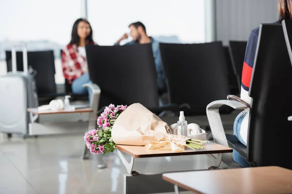 Bouquet enveloppé de fleurs sur la table près de la bouteille avec désinfectant et écouteurs sans fil avec des personnes interracial sur fond flou — Photo de stock