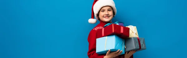 Plano panorámico de niño feliz en sombrero de santa celebración de regalos aislados en azul - foto de stock