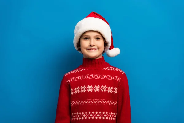 Glücklicher Junge mit Weihnachtsmütze und rotem Pullover, der auf blauem Grund in die Kamera blickt — Stockfoto