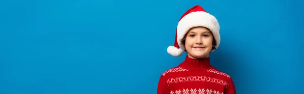 Panoramic crop of joyful boy in santa hat and red sweater looking at camera on blue — Stock Photo