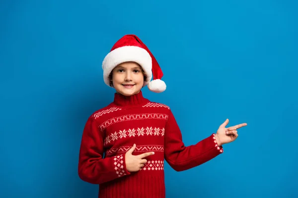 Menino alegre em chapéu de santa e suéter vermelho apontando com os dedos no azul — Fotografia de Stock