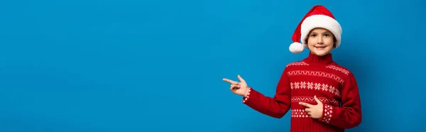 Panoramic concept of joyful boy in santa hat and red sweater pointing with fingers on blue — Stock Photo