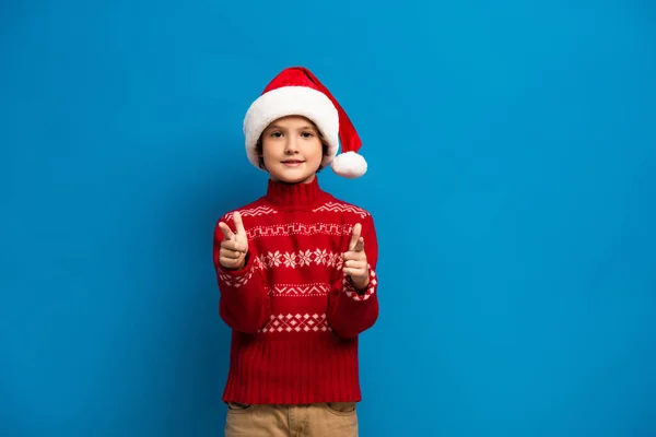 Joyeux garçon en santa chapeau et pull rouge pointant avec les doigts tout en regardant la caméra sur bleu — Photo de stock