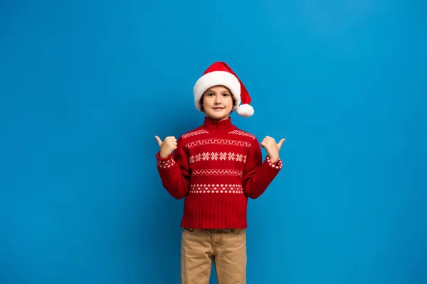 Joyful kid in santa hat and red sweater showing thumbs up while looking at camera on blue — Stock Photo