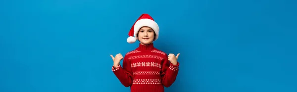 Panoramaaufnahme eines fröhlichen Kindes mit Weihnachtsmannmütze und rotem Pullover, das die Daumen nach oben zeigt, während es isoliert auf blau in die Kamera blickt — Stockfoto