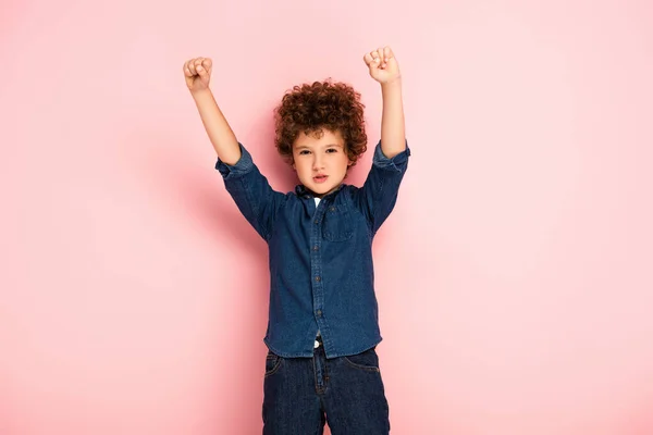 Excited curly boy with hands above head and clenched fists showing winner gesture on pink — Stock Photo