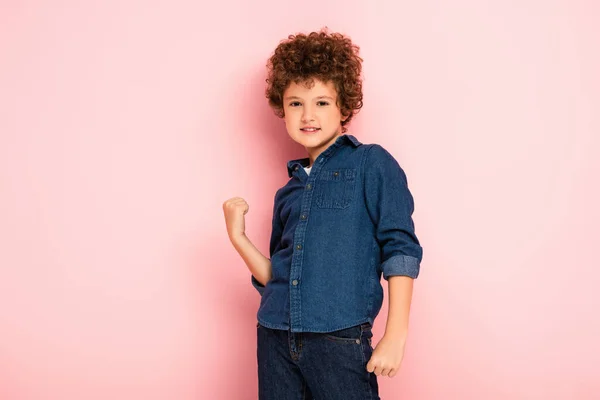 Pleased and curly boy with clenched fsts looking at camera on pink — стоковое фото
