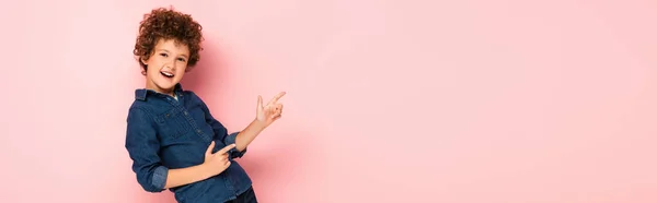 Panoramic concept of excited curly boy in denim shirt pointing with fingers on pink — Stock Photo