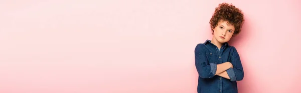 Panoramic crop of serious and curly boy standing with crossed arms on pink — Stock Photo