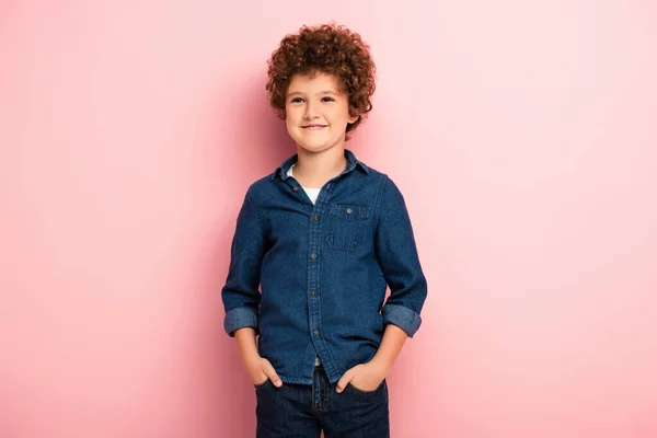 Довольный и вьющийся мальчик в джинсовой рубашке, стоящий с руками в карманах на розовом — стоковое фото