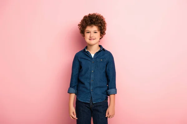 Веселый и кудрявый мальчик в джинсовой рубашке, стоящий на розовом — стоковое фото