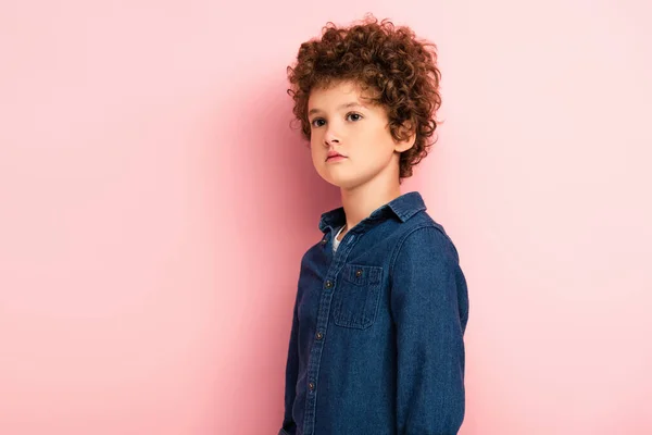 Niño rizado en camisa de mezclilla de pie y mirando hacia otro lado en rosa - foto de stock