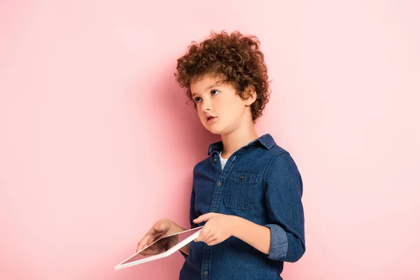 Скептически настроенный и кудрявый ребенок держит цифровой планшет на розовом — стоковое фото