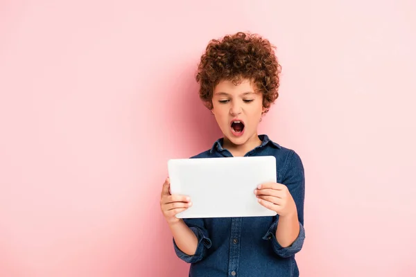 Garoto irritado e encaracolado gritando e olhando para tablet digital em rosa — Fotografia de Stock