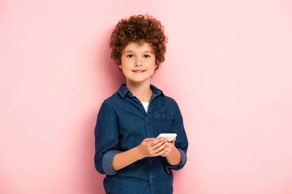 Pleased boy in denim shirt holding smartphone on pink — Stock Photo
