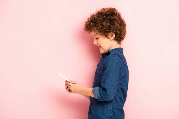 Side view of pleased boy in denim shirt holding smartphone on pink — Stock Photo
