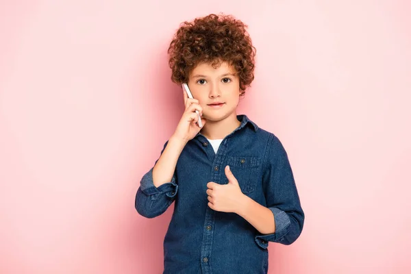 Niño rizado en camisa de mezclilla hablando en el teléfono inteligente y mostrando el pulgar hacia arriba en rosa - foto de stock