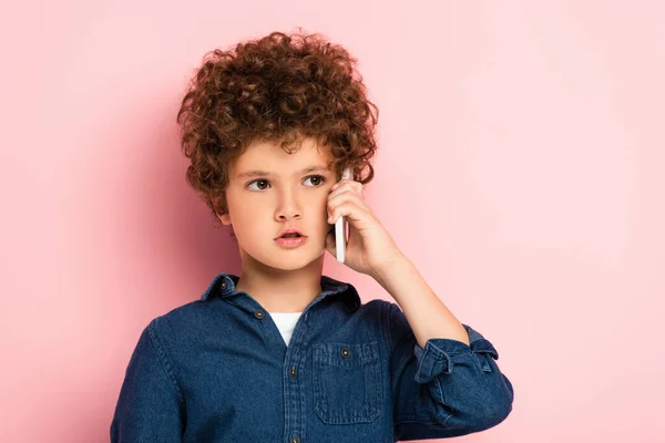 Menino encaracolado em camisa jeans falando no telefone celular e olhando para longe em rosa — Fotografia de Stock