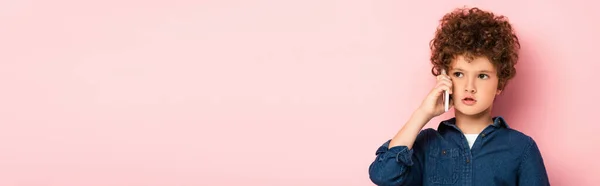 Horizontal image of curly boy in denim shirt talking on mobile phone and looking away on pink — Stock Photo