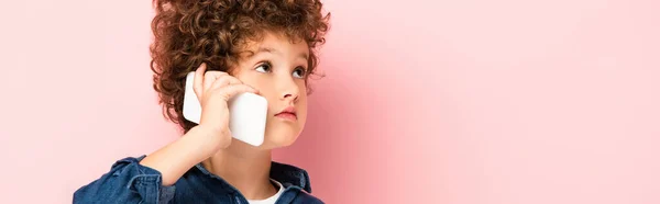 Conceito horizontal de menino encaracolado na camisa jeans falando no telefone celular e olhando para longe em rosa — Fotografia de Stock
