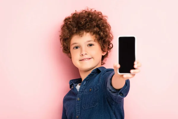 Foco seletivo do menino encaracolado na camisa jeans segurando smartphone com tela em branco no rosa — Fotografia de Stock