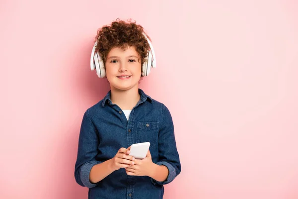 Niño alegre y rizado escuchando música en auriculares inalámbricos y sosteniendo el teléfono inteligente en rosa - foto de stock