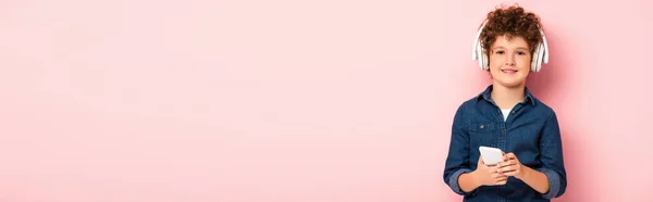 Панорамный урожай радостного мальчика, слушающего музыку в беспроводных наушниках и держащего смартфон на розовом — стоковое фото