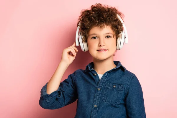 Niño alegre y rizado escuchando música y tocando auriculares inalámbricos en rosa - foto de stock