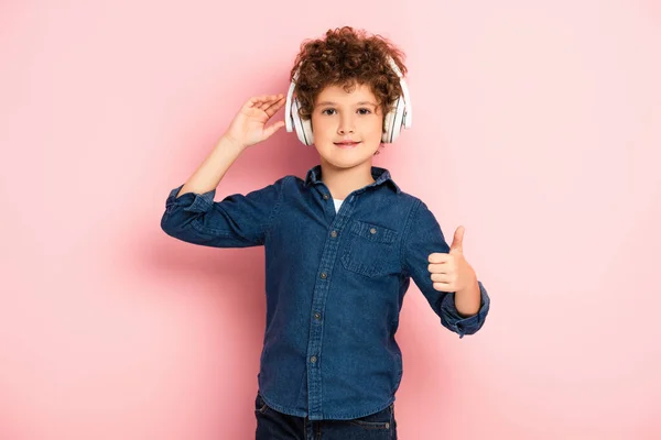 Niño alegre y rizado escuchando música en auriculares inalámbricos mientras muestra el pulgar hacia arriba en rosa - foto de stock