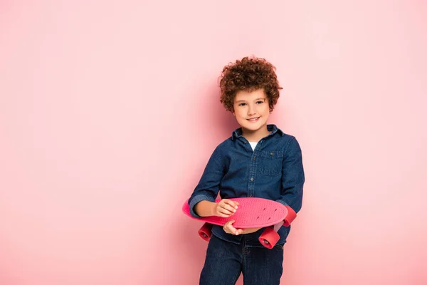 Zufriedener Junge hält Penny-Board und lächelt auf rosa — Stockfoto