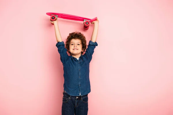 Curly boy holding penny board above head on pink — Stock Photo