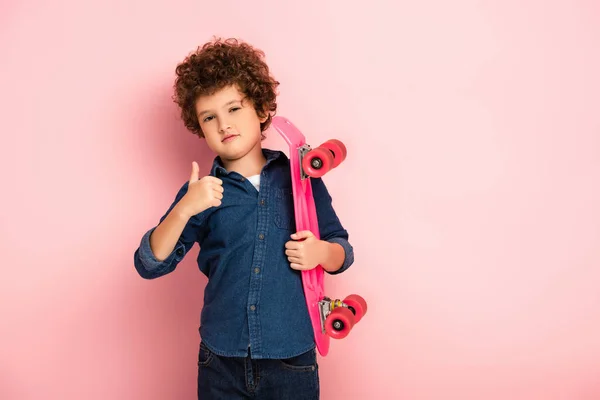 Curly boy holding penny board and showing thumb up on pink — Stock Photo