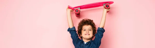 Panoramic crop of curly boy holding penny board above head on pink — Stock Photo