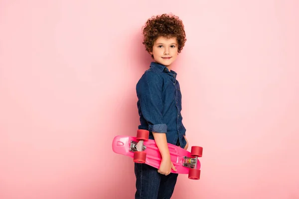 Riccio ragazzo tenendo penny board e sorridente su rosa — Foto stock