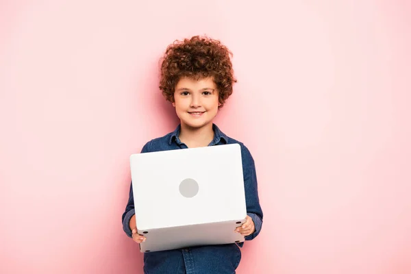 Веселый и кудрявый мальчик держит ноутбук на розовом — стоковое фото