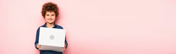 Concept panoramique de garçon joyeux et bouclé tenant ordinateur portable sur rose — Photo de stock