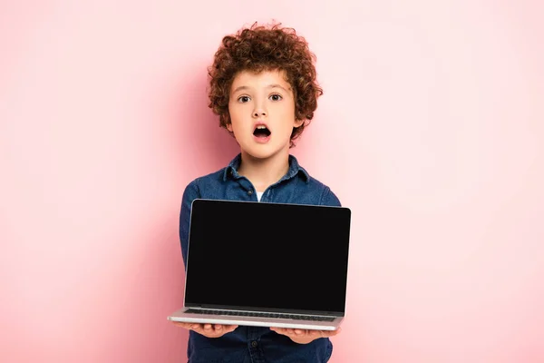 Menino chocado e encaracolado segurando laptop com tela em branco no rosa — Fotografia de Stock